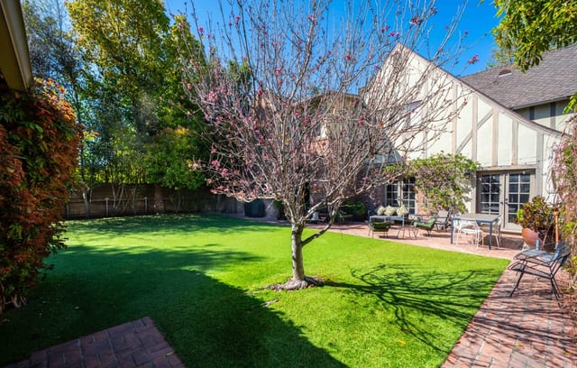 Level lawn enclosed by fencing and mature trees, with a blossoming tree at its centre and garden furniture along the far edge.