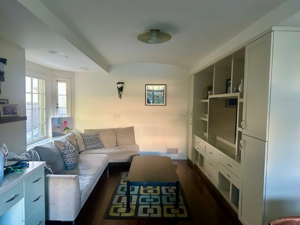 Narrow den with a pale sectional sofa, a long low table on a patterned rug, fitted white cabinetry and open shelving along one wall, and a glazed door to the garden.
