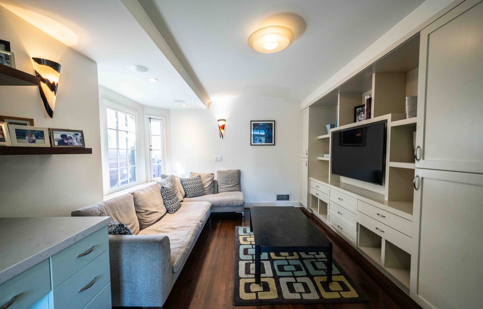 Narrow den with a long built-in sofa, fitted cabinetry and shelving, a wall-mounted television and a patterned rug.