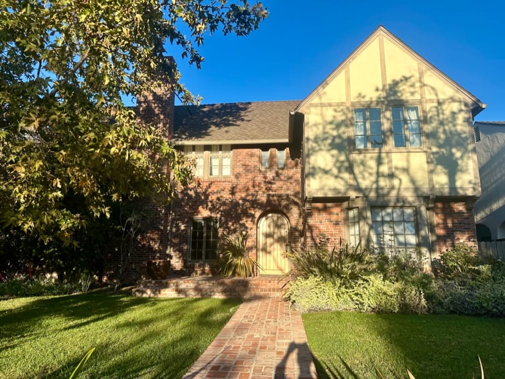 Front elevation of a brick and half-timber Tudor house with a steeply pitched gable, a tall brick chimney, leaded upper windows and an arched wooden front door, reached by a brick path across a level front lawn, with a mature tree overhanging from the left.