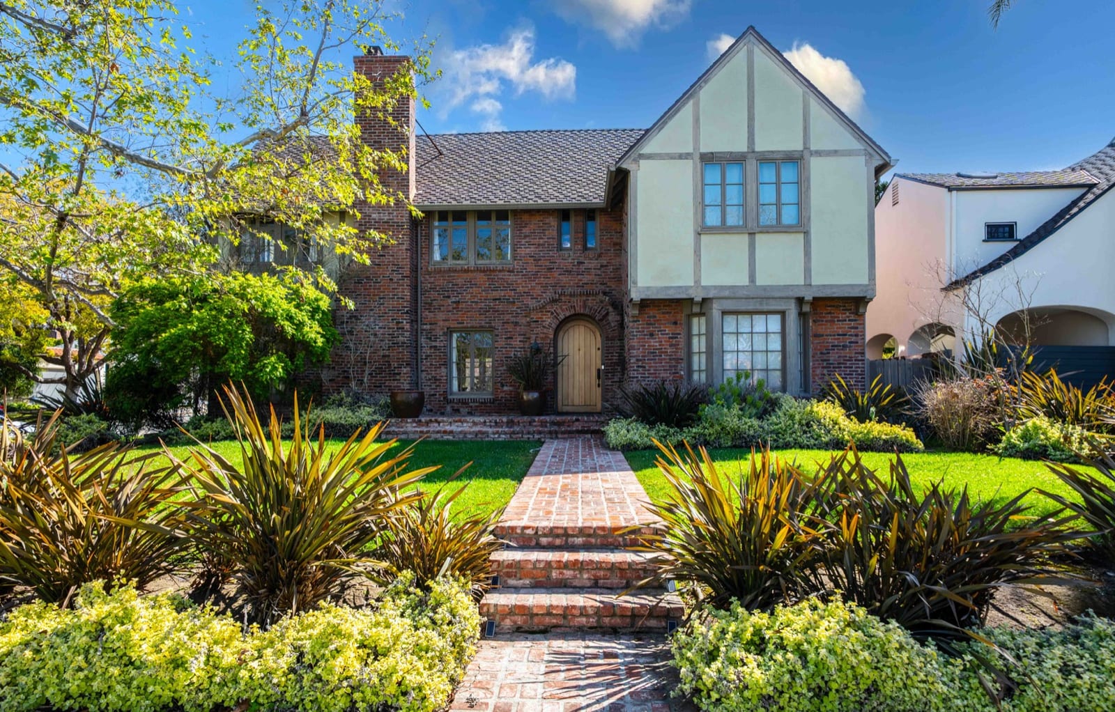 Front elevation of a brick and half-timber Tudor house behind a mature planted garden, with a brick path leading to an arched front door.