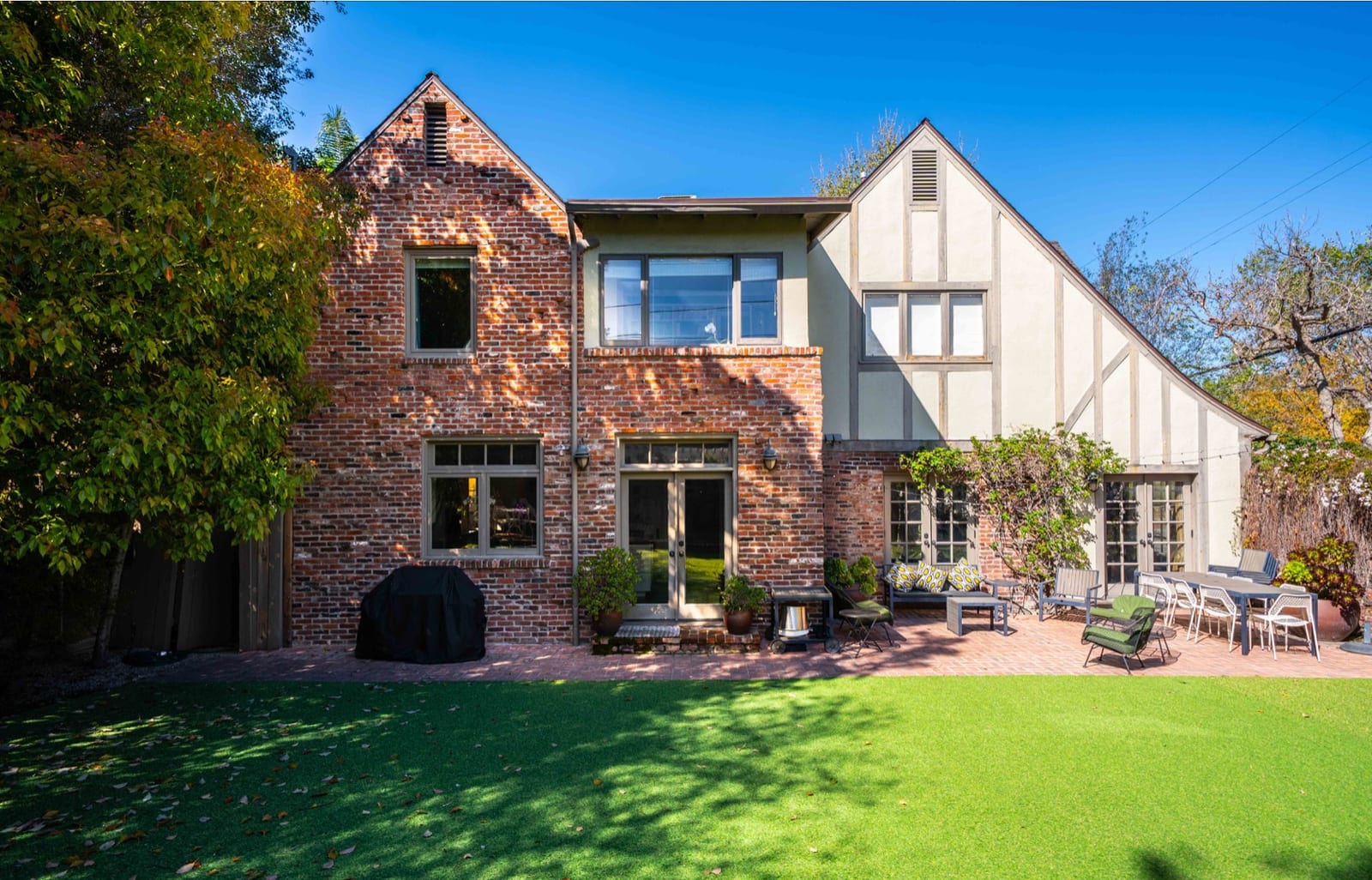 Rear elevation of the Tudor house in brick and half-timber, with tall gables and climbing planting, seen across an open lawn.