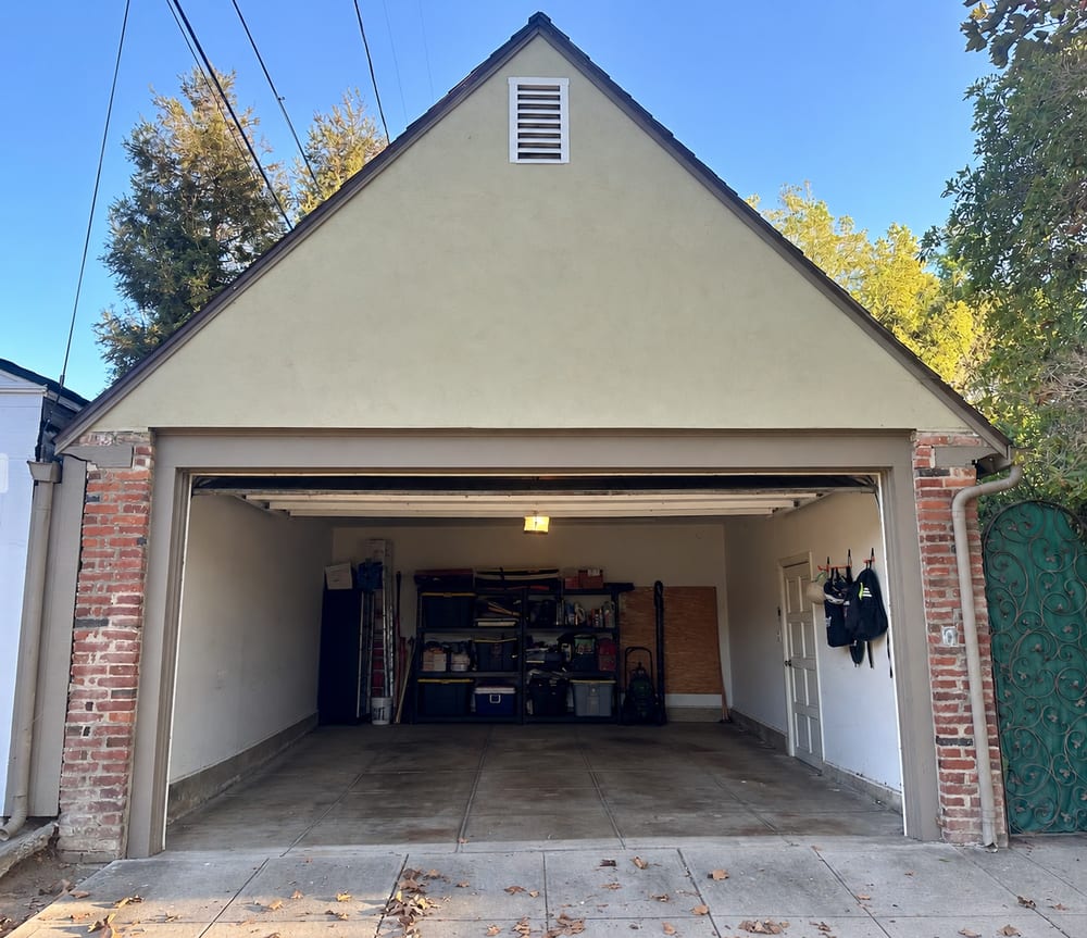 Detached garage with a stuccoed gable end and brick piers, its door open on a clear two-car bay with a concrete floor and storage shelving along the back wall.