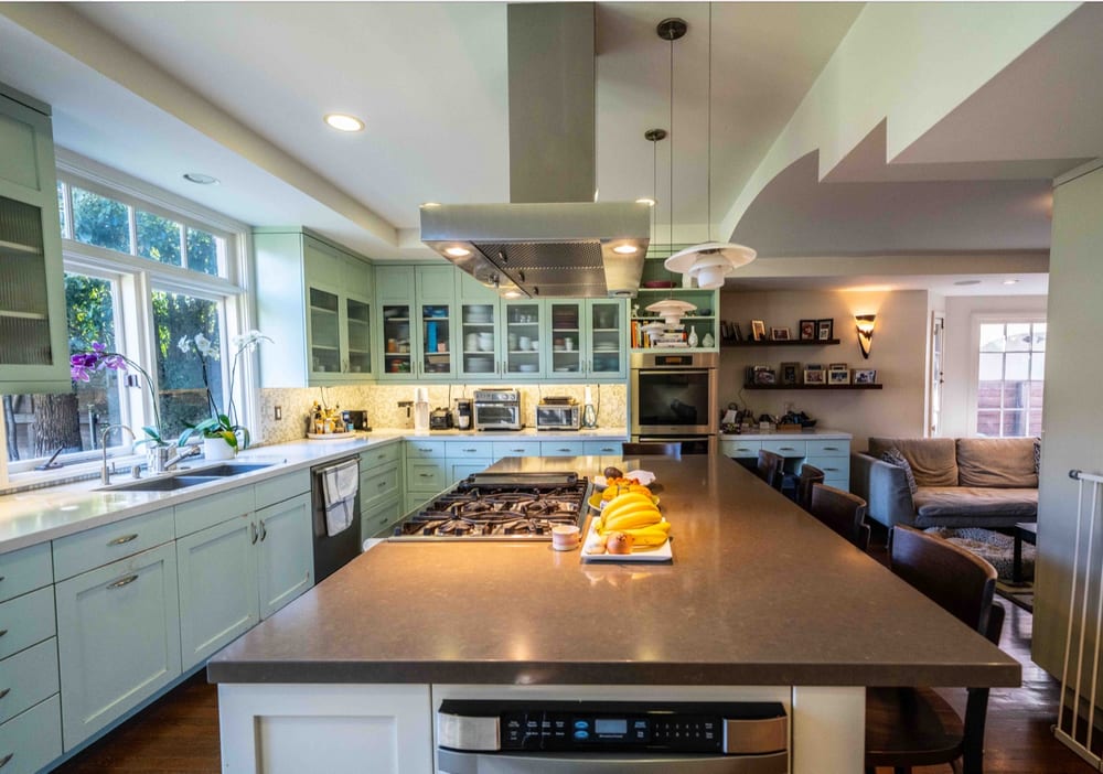 Open kitchen with pale green cabinetry, a long island with a range and extraction hood, and a raised ceiling opening onto a seating area.