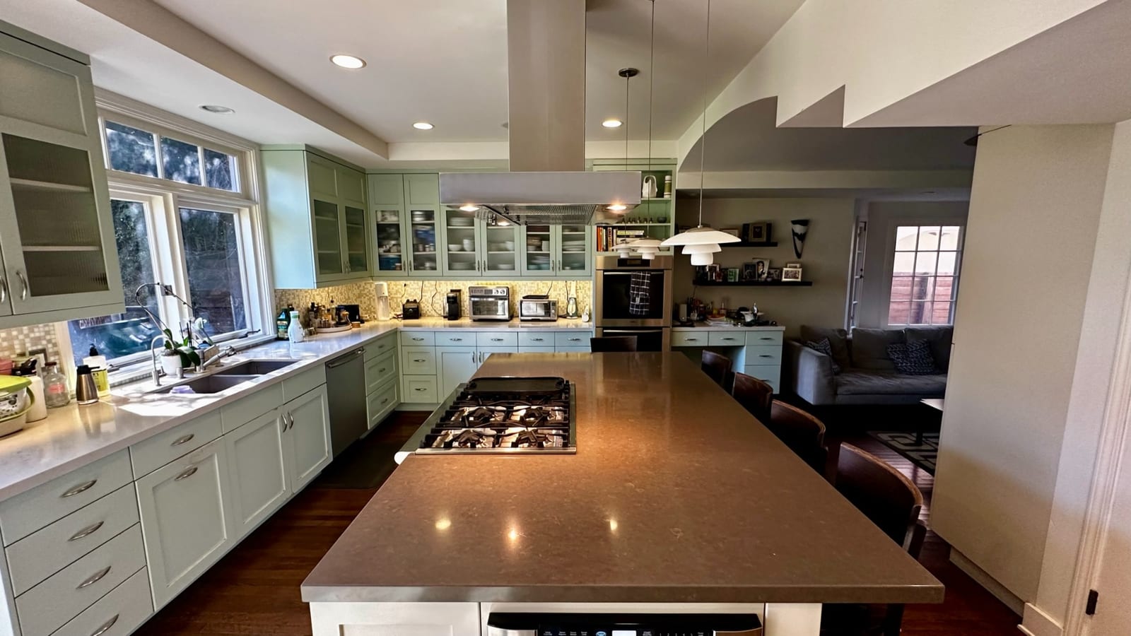 Kitchen with pale green cabinetry and glass-fronted upper cupboards, a long stone-topped island with a gas range and an extractor hood above it, opening past a built-in desk to a sitting area with a sofa.