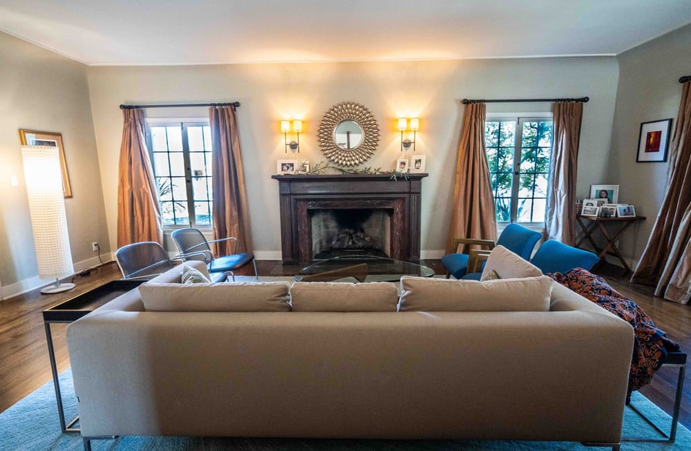 Living room with a carved stone fireplace flanked by wall sconces, tall curtained windows either side and low seating facing the hearth.