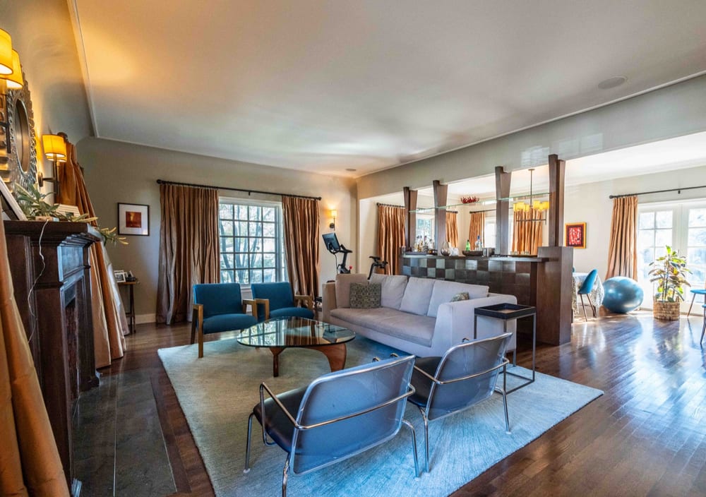 Wider view of the living room showing dark hardwood floors, an area rug, armchairs and a columned opening through to the next room.
