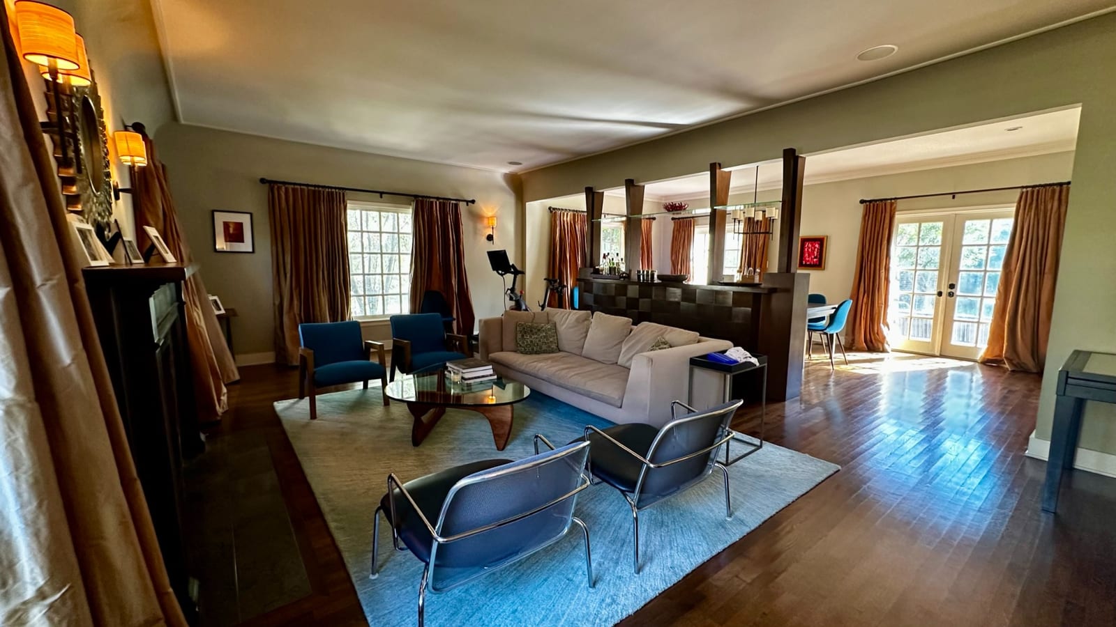 Wide view of the living room looking past the fireplace and seating area to a dark wood bar and a dining table beyond, with tall curtained windows along one wall and French doors opening to the garden at the far end.