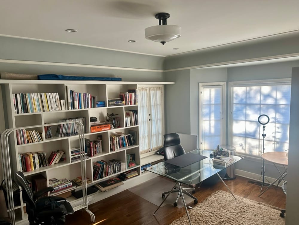 Downstairs office in pale blue with floor-to-ceiling built-in bookshelves, a glass-topped desk and a black desk chair, lit by a bay of tall windows.