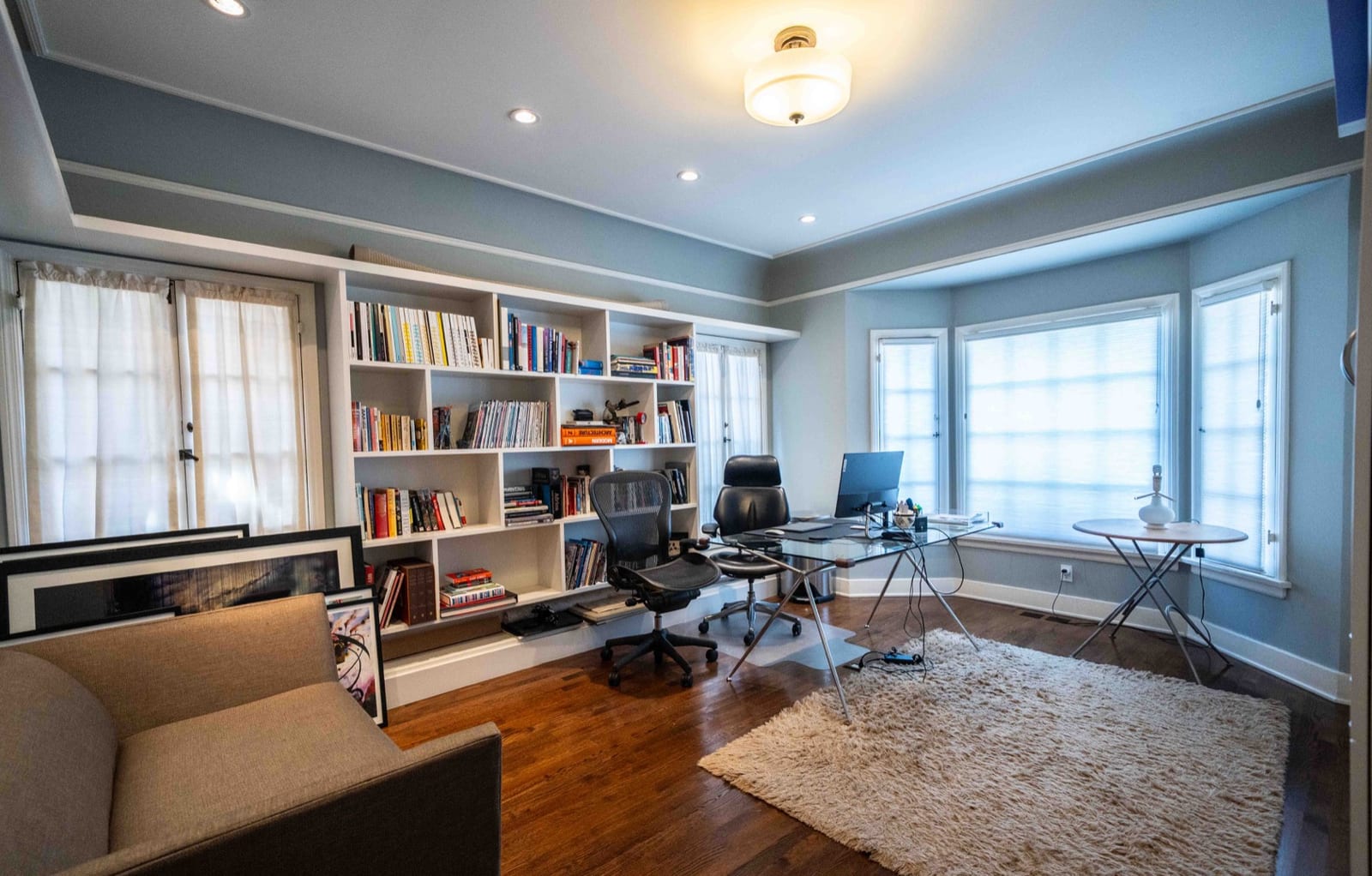 Downstairs office with pale blue walls, built-in bookshelves, a desk in a bay window and a glass-topped table.