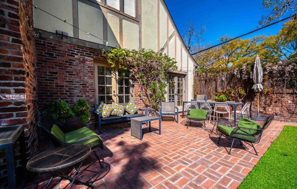 Brick-paved patio against the house with outdoor armchairs, a low table, a dining set and a parasol beneath climbing greenery.
